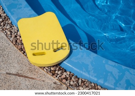 A yellow swim board on beside of the swimming pool. Swim board are a flotation aid used to develop a swimmer's kicking action. Royalty-Free Stock Photo #2300417021