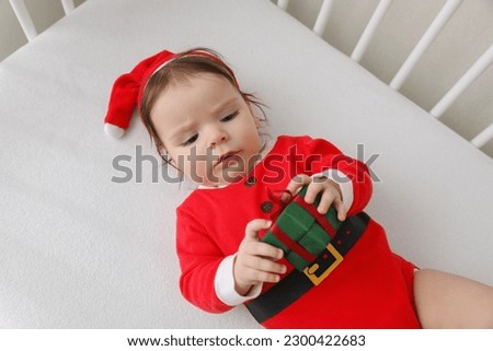 Cute baby wearing festive Christmas costume with gift box in crib, above view