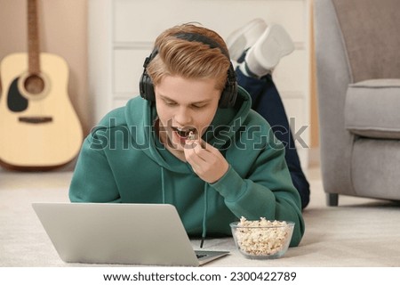 Teenage boy with headphones eating popcorn while using laptop at home
