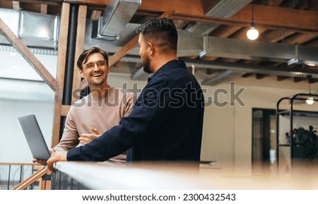 Two business men talking to each other about their tech work. Business colleagues discussing efficient ideas to finish a project. Male professionals working in a startup. Royalty-Free Stock Photo #2300432543