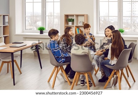 Group of elementary school children and their teacher discussing something while sitting in a circle on comfortable chairs in a modern classroom all together