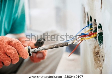 Close up of man in work gloves using electric repair solder welding tool while installing electrical cables in apartment under renovation. Male electrician working with electric heater soldering iron. Royalty-Free Stock Photo #2300460107