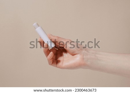 Female hand holding white hygienic lipstick on beige isolated background. Weather protection, skin care and beauty brand product concept Royalty-Free Stock Photo #2300463673