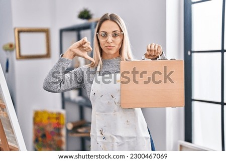 Young blonde painter woman holding wooden case at art studio with angry face, negative sign showing dislike with thumbs down, rejection concept 