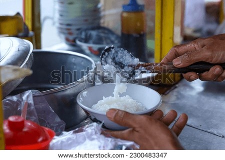 Chicken porridge seller is preparing order for customers. Bubur Ayam (Chicken Porridge), Indonesian Traditional Food. Hand in frame Royalty-Free Stock Photo #2300483347