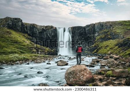 Beautiful view of Gufufoss waterfall flowing with young asian tourist woman standing on the rock in summer at Seydisfjordur, East of Iceland