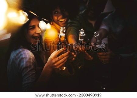 Happy friends enjoying night party on rooftop terrace - Group of multiracial young people drinking beer bottle at brewery pub garden - Life style concept with guys and girls hanging outdoors