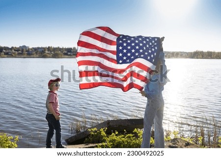 woman and child are standing on shore of lake holding an American flag waving in hands, beautifully illuminated by the sun. patriotic holiday, Independence Day. Pride to be an American.