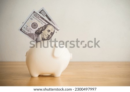 Piggy bank and US dollar banknotes on wooden table with white wall background copy space. Personal finance, money savings management for education, retirement, investment, travel, etc concept.