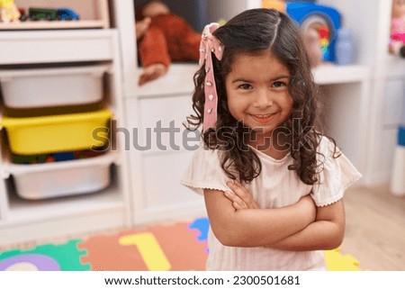 Adorable hispanic girl smiling confident sitting on floor with arms crossed gesture at kindergarten