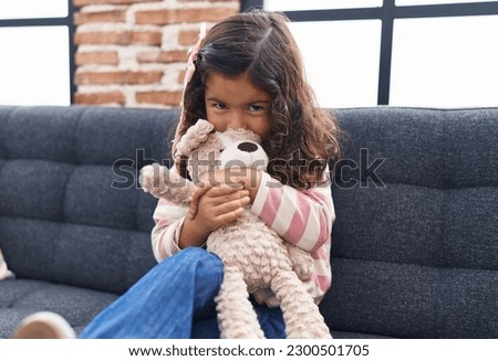 Adorable hispanic girl kissing and hugging teddy bear sitting on sofa at home