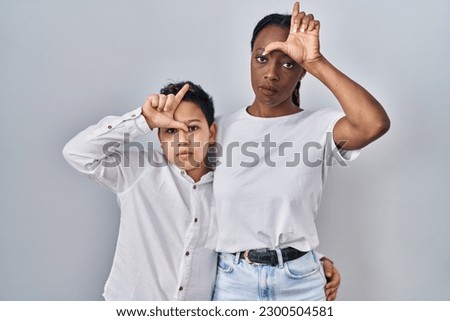 Young mother and son standing together over white background making fun of people with fingers on forehead doing loser gesture mocking and insulting. 