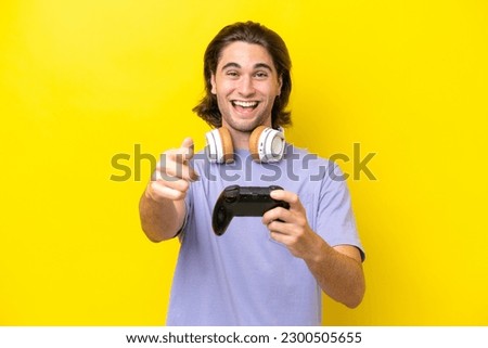 Young handsome caucasian man playing with a video game controller over isolated on yellow background surprised and pointing front