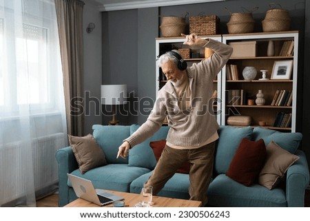 Happy senior old grandfather man in wireless headphones dancing, singing at home, choosing favorite energetic disco music in mobile application, entertaining indoors. Retired people relax activities Royalty-Free Stock Photo #2300506243