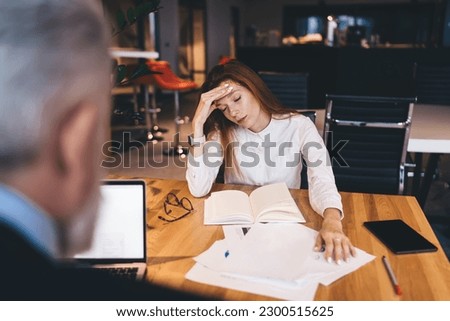 Exhausted female entrepreneur in formal attire sitting at wooden table with papers and planner and touching head while male coworker using laptop during workday in modern business center