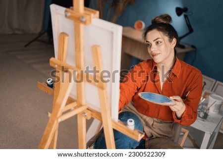 side view focused ethnic woman in casual wear sits on chair draws on canvas with colorful paints and brush in small room. Makes a stroke of paint on canvas.