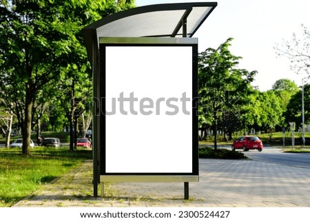 bus shelter with blank ad panel. billboard display. empty white lightbox sign at bus stop. glass structure. city transit station. urban street. green park setting. outdoor bus shelter advertising