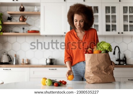 Happy Young Black Lady Unpacking Paper Bag With Groceries After Food Shopping, Smiling Millennial African American Woman Putting Fresh Vegetables And Fruits On Table, Standing In Kitchen At Home Royalty-Free Stock Photo #2300535227
