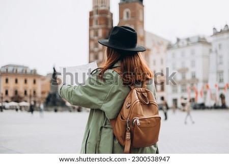 Attractive young female tourist is exploring new city. Redhead girl walking and holding a paper map in Krakow. Traveling Europe in spring. Urban lifestyle banner, Back view