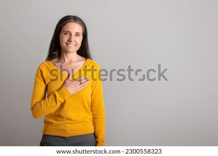 Cheerful pretty caucasian elderly lady puts her hand to chest, make thanks gesture on gray studio background, copy space. Great news, thanks sign, ad and offer, people emotions