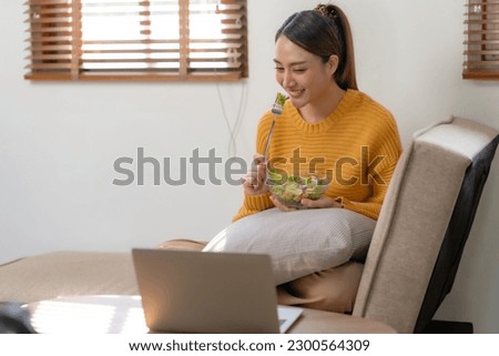 Happy young Asian woman eating salad at home and using laptop computer.