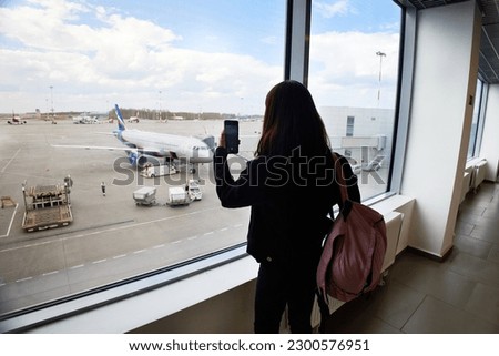 A young girl at the airport looks out the window and takes pictures of planes. View from the back. The concept of travel. 