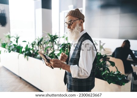 Side view of elderly male in eyeglasses and hat using mobile phone in hands with notepad while looking at screen and sending message while standing in blurred modern office
