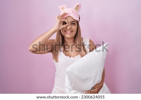 Young blonde woman wearing pyjama hugging pillow doing ok gesture with hand smiling, eye looking through fingers with happy face. 