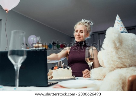 Young woman in party cap drinks champagne and eating cake, having online birthday party with friends. Girl speaking, making video chat, lifestyle blog vlog, webcam view. Stay home, quarantine concept.