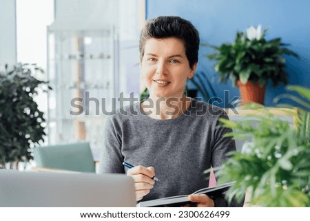Smiling neutral gender middle-aged person making notes in her paper notebook, using laptop while working indoors in her workstation in an open space office. Online video call, IT HR, recruiting. Royalty-Free Stock Photo #2300626499