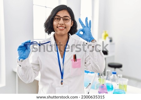 Young hispanic woman working at scientist laboratory holding safety glasses doing ok sign with fingers, smiling friendly gesturing excellent symbol 