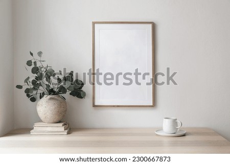 Empty wooden picture frame, poster mockup hanging on beige wall background. Vase with green eucalyptus tree branches on table. Cup of coffee, books. Working space, home office. Modern art display. 