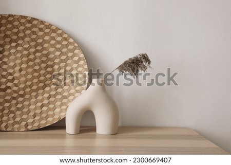 Boho ethnic home decor. Elegant neutral still life with modern vase and  dry grass. Decorative checkered  rattan tray, plate on wooden table. Empty beige wall, copyspace, no people. Royalty-Free Stock Photo #2300669407