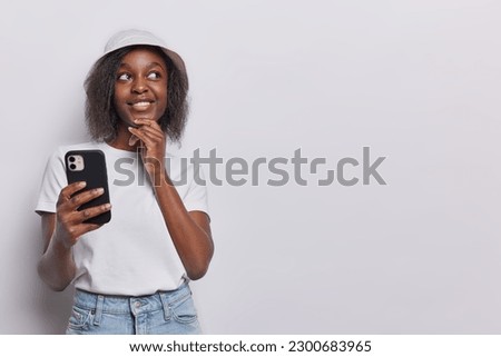 Horizontal shot of thoughtful dark skinned woman keeps hand on chin concentrated aside downloads special app for chatting searches suitable tour for spending summer vacation dressed in casual clothing