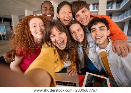 Selfie with mobile phone of multiracial cheerful group of students gathered in hall of university building. Happy young friends hugging laughing and posing for photo. People smiling on campus.
