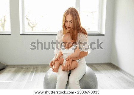Happy mother holding baby girl while bouncing on fitness ball in cozy home. Stability ball exercises for neurodevelopment of baby. Royalty-Free Stock Photo #2300693033