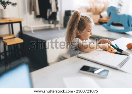 Child with ponytail sitting at table expressing creativity while drawing in sketchbook with multicolored markers while spending time at home