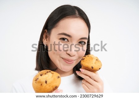 Image of korean woman enjoys eating baked pastry, showing two tasty cupcakes near face and smiles, white background.