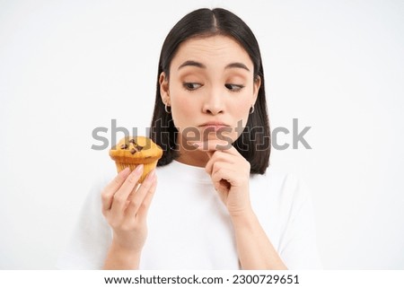 Image of pensive korean woman, looks at cupcake with thoughtful face, thinking about dessert, white background.