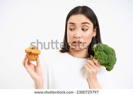 Image of vegeterian girl, avoids pastry, sticks to broccoli, eating vegetables instead of cupcake, white background.