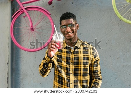 Carefree african american guy in casual wear holding tropical cocktail urban summer background. Cool young man drinking yummy alcohol-free beverage on summer vacation