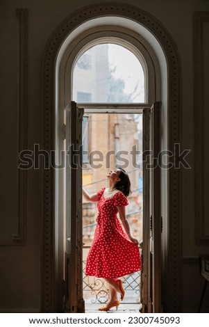 a beautiful and happy brunette women in red retro dress and gloved in the balcony doorway . the concept of the stylist's work. thematic photo shoot.