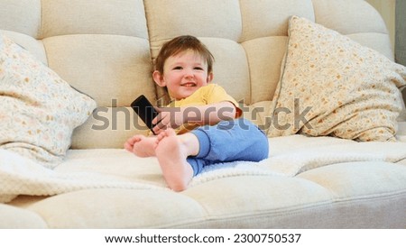 The little child sits on the sofa, watching TV with a remote control in hand and a smile on their face. The happy person is watching their favorite show on TV, comfortably seated on the beige sofa.