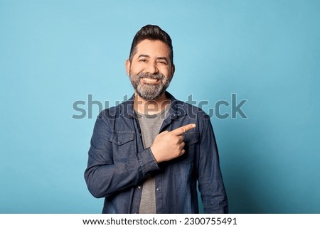 Mid adult latin man smiling pointing to the side showing copy blank space, isolated over blue background. Indoor studio shot. Advertisement, presentation concept. Copy space. Royalty-Free Stock Photo #2300755491