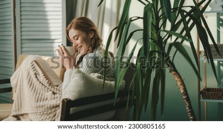Young relaxed woman in casual outfit reading book and drinking tea while relaxing on sofa in cozy living room at home. Enjoying free time on weekend at home Royalty-Free Stock Photo #2300805165