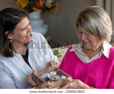 Daughter giving a gift to her mother on Mother's Day. Gesture of love and affection for a special person.
Box of chocolates and cookies. she wears a fuchsia sweater and the daughter a white shirt.