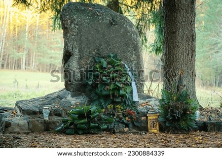 Gravestone standing tall in cemetery with big flower covering near tree with bright sunny meadow on the background
