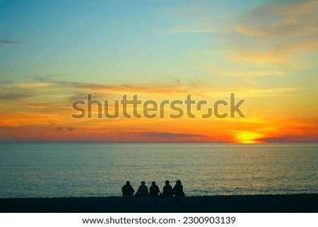 Young teenage people - guys and girls - sit on the beach and watch the sunset. Group of five happy people sits on background of empty sunset beach. Travel or sea vacations concept. back, rear view. 