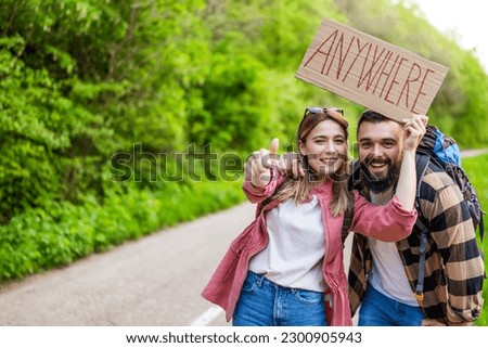 Happy couple hitchhiking on roadside trying to stop car. They are holding cardboard with inscription. Royalty-Free Stock Photo #2300905943