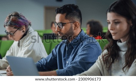 Young Handsome Male Student Studying in University with Diverse Multiethnic Classmates. Young Man is Writing Down Lecture Summary in Notebook to Prepare for College Exams Royalty-Free Stock Photo #2300906173